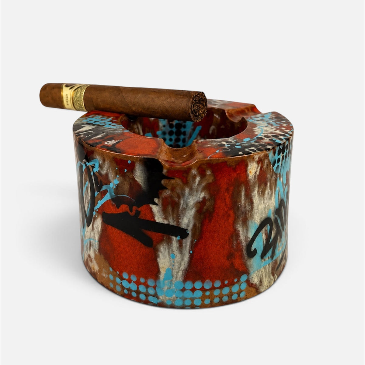 A cylindrical ceramic ashtray with a distressed, graffiti-style pattern in red, brown, black, and turquoise blue, featuring a single cigar resting horizontally across the top opening, set against a plain light gray background.