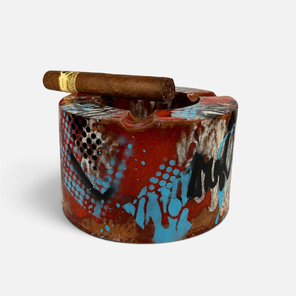 A cylindrical ashtray with a graffiti-style design in red, blue, black, and brown, featuring a brown cigar resting across the top, set against a plain light gray background.