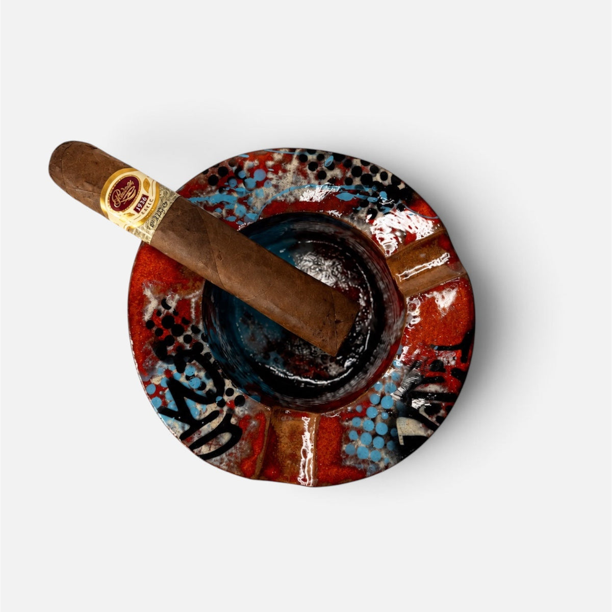 A top-down view of a cigar resting on a round, hand-painted ceramic ashtray. The ashtray features a vibrant red, blue, and black pattern with a small gold chain accent.