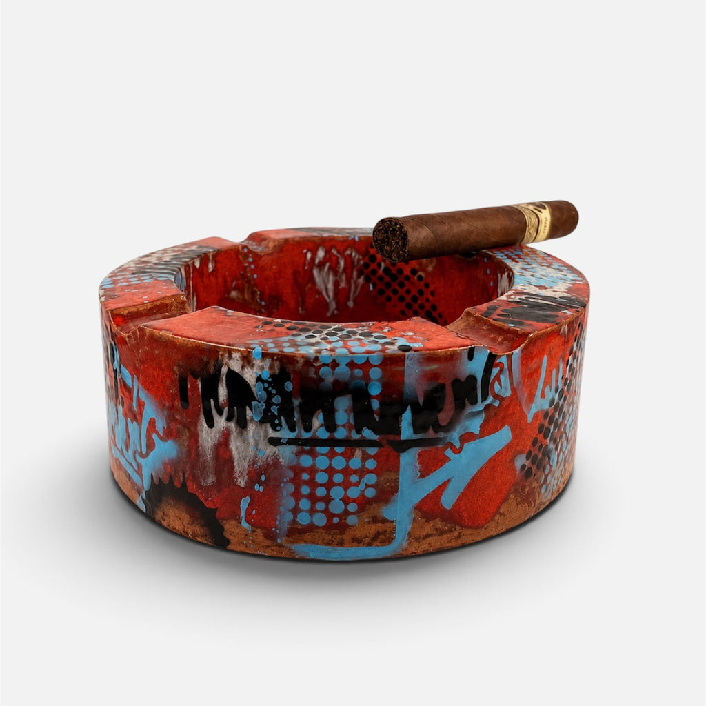 A cylindrical ceramic ashtray with a vibrant graffiti-style pattern in red, orange, blue, and black, featuring a brown cigar resting on top, set against a plain light gray background.