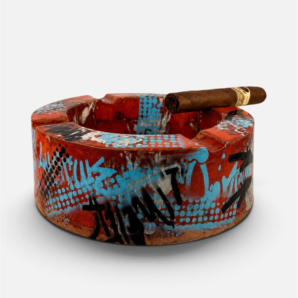 A circular ceramic ashtray with a vibrant graffiti-style design in red, blue, black, and white, featuring a brown cigar resting on top, set against a plain light gray background.