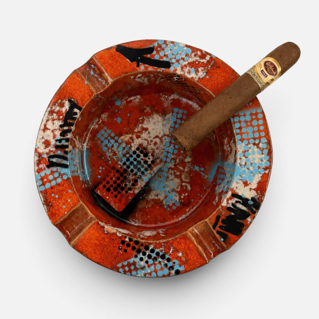 A top-down view of a ceramic ashtray with a cigar resting inside, set against a plain light gray background. The ashtray features a vibrant orange and red pattern with blue and black accents, and the cigar band displays the 'Cigars Corona' logo.