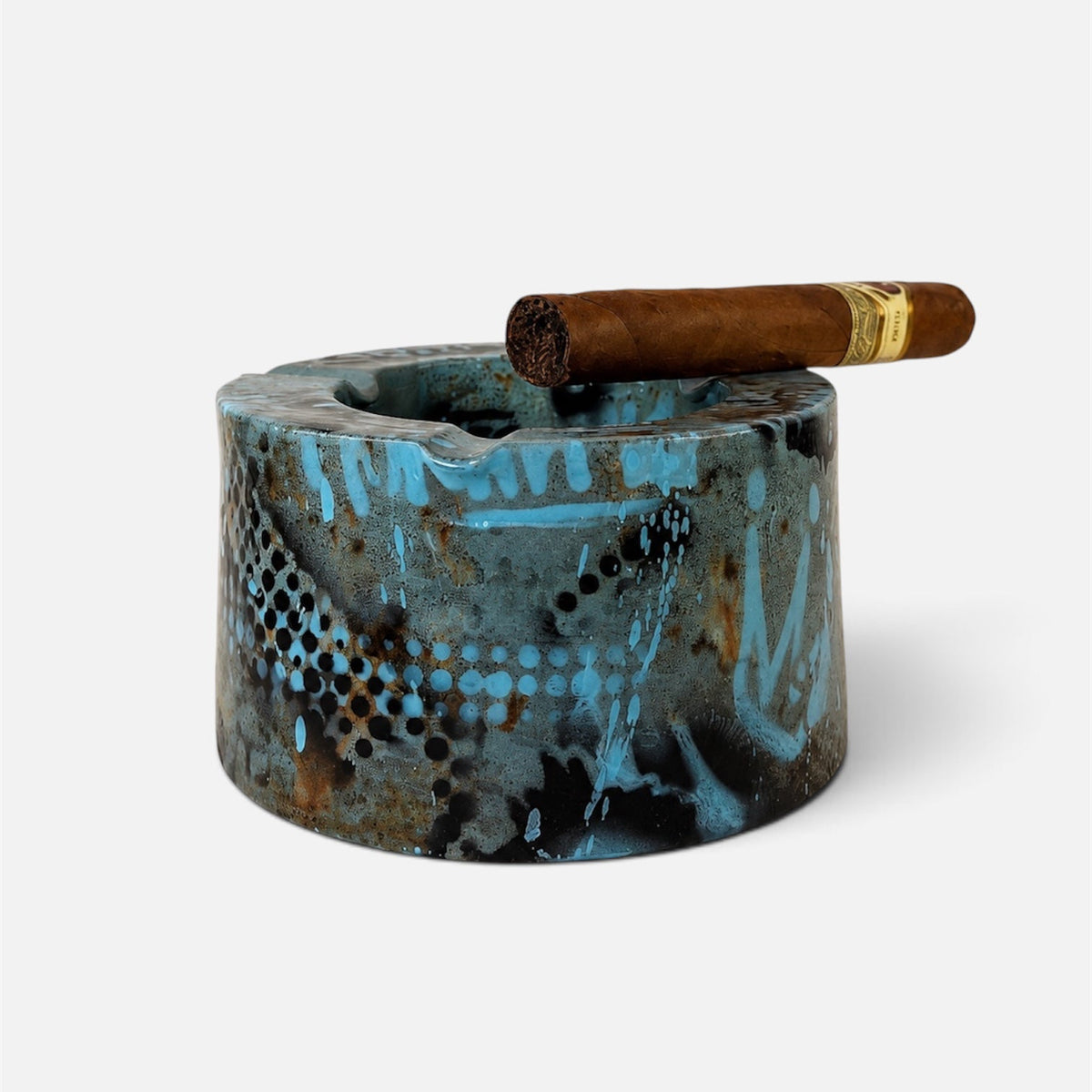 A cylindrical ceramic ashtray with a textured, distressed finish in shades of blue, brown, and black, featuring a white painted graffiti-style pattern. A single brown cigar rests horizontally across the rim of the ashtray.
