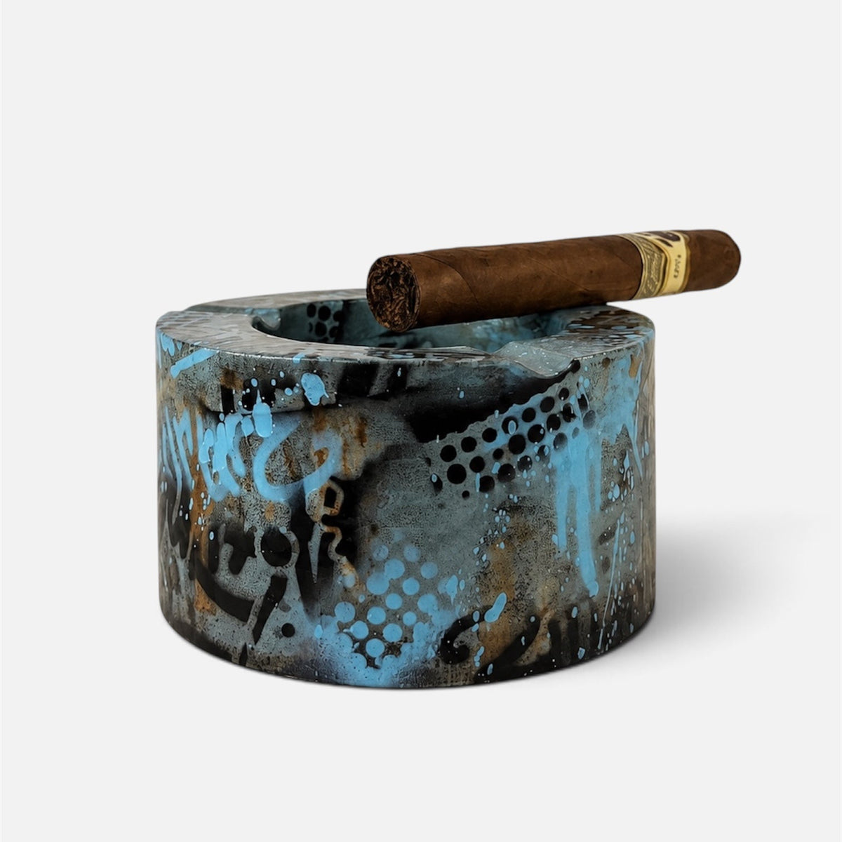A cylindrical smoking ashtray with a distressed blue, black, and brown abstract pattern, shown with a brown cigar resting across the top rim on a light gray background.