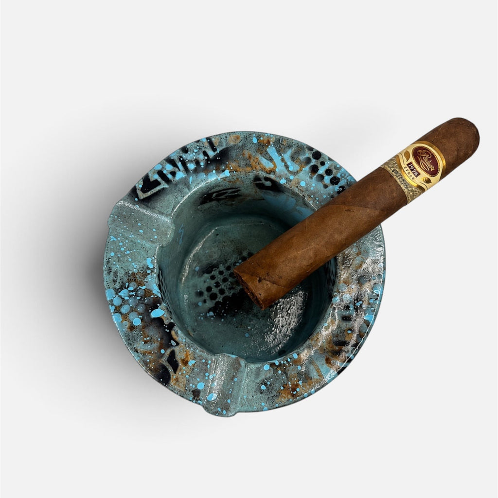 A top-down view of a distressed turquoise blue ceramic ashtray with a brown cigar resting inside, set against a plain light gray background.