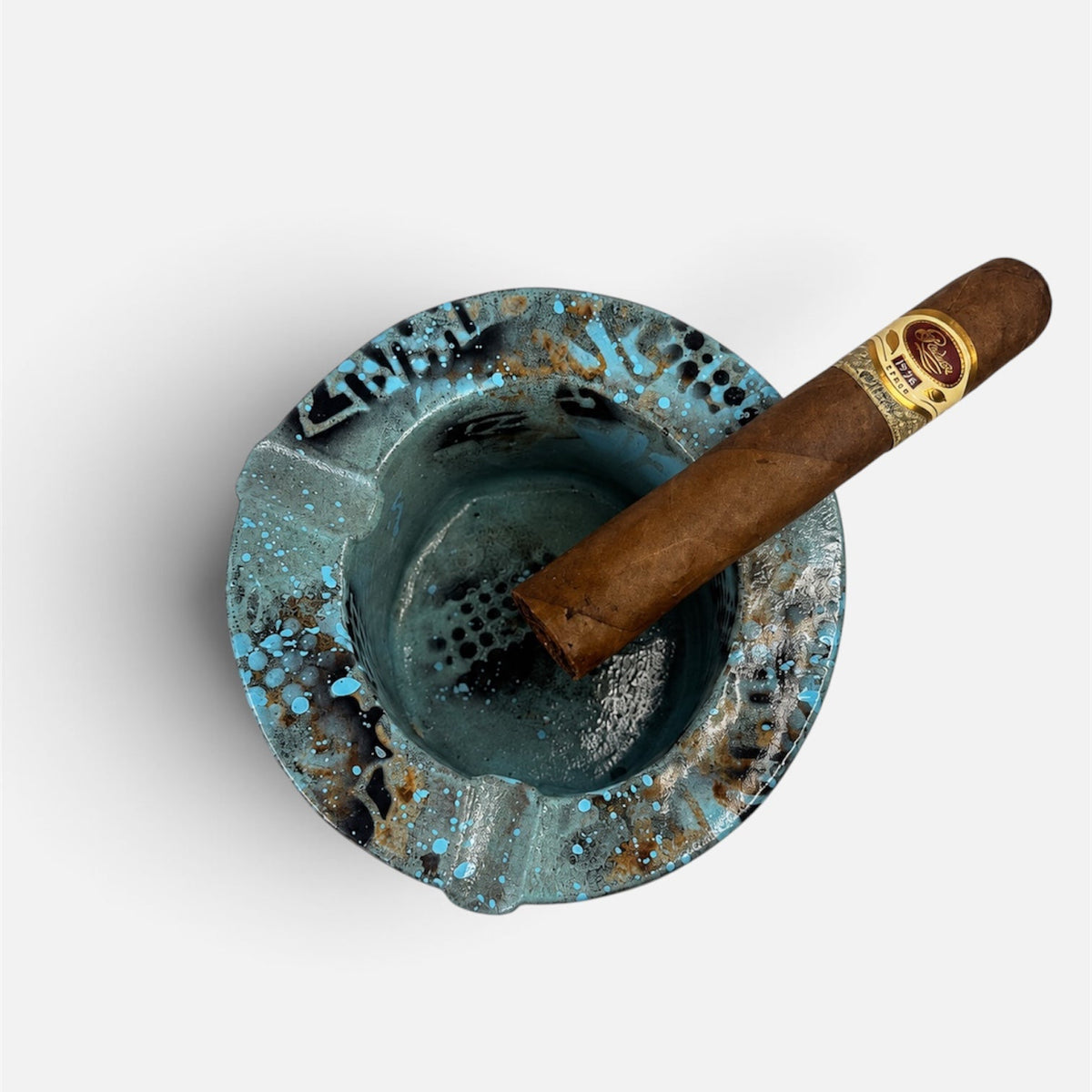 A top-down view of a distressed turquoise blue ceramic ashtray with a brown cigar resting inside, set against a plain light gray background.