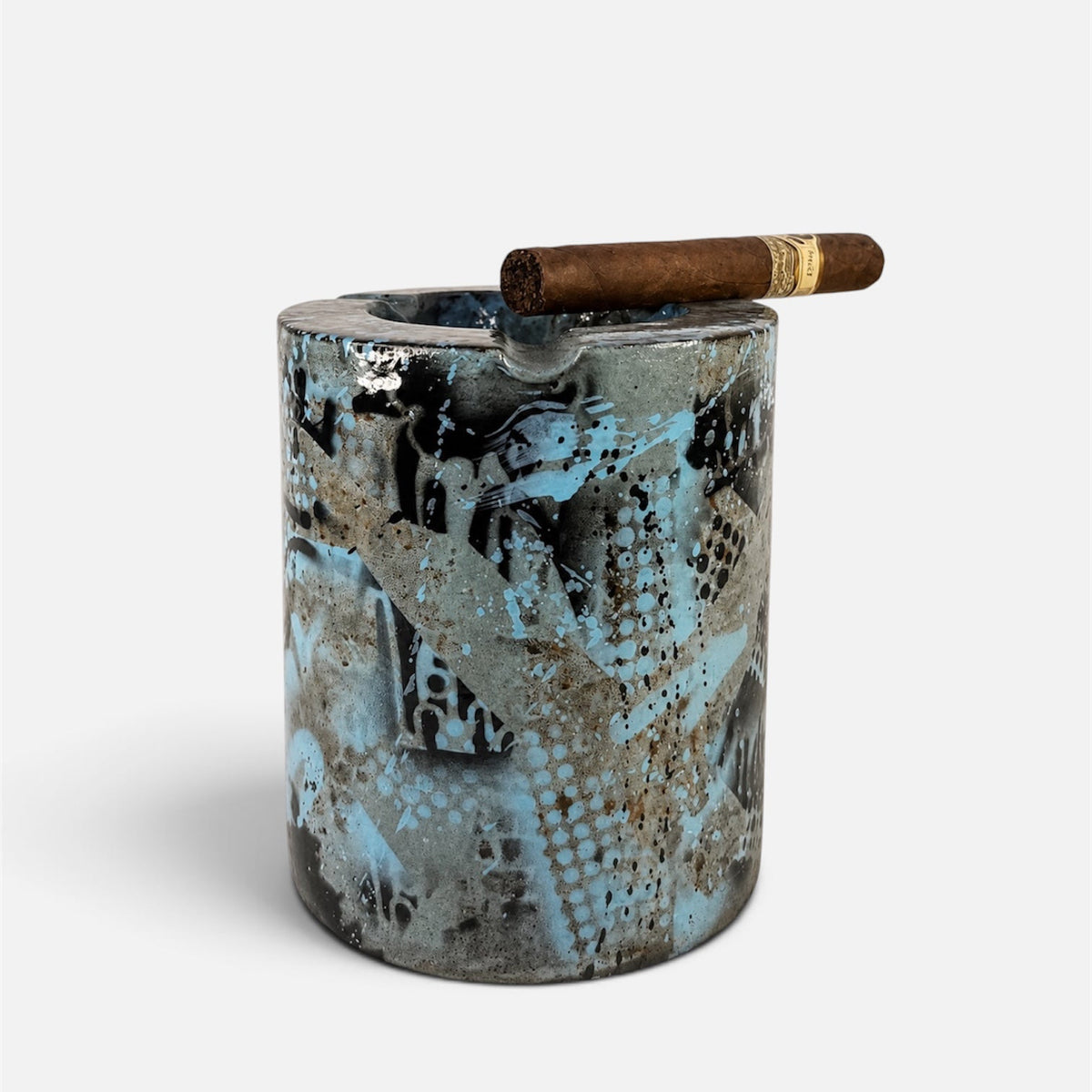 A cylindrical ceramic cigar ashtray with a textured, abstract pattern in blue, black, and gray, featuring a brown cigar resting across the top rim, set against a plain light gray background.