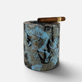A cylindrical ceramic cigar ashtray with a textured, distressed pattern in black, gray, and light blue, featuring a brown cigar resting on top against a light gray background.