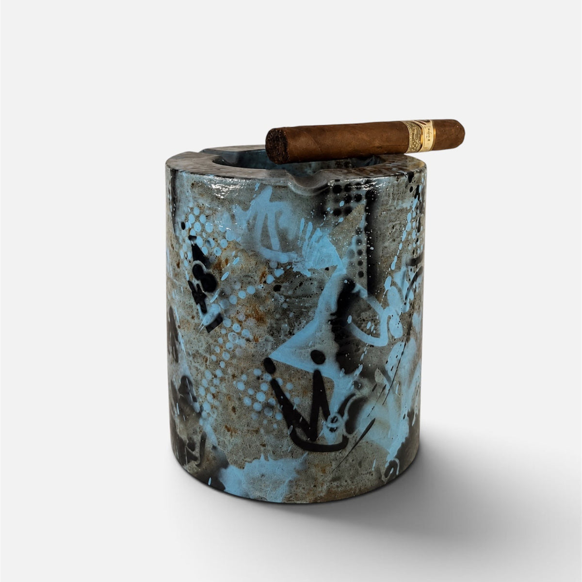 A cylindrical ceramic cigar ashtray with a textured, distressed pattern in black, gray, and light blue, featuring a brown cigar resting on top against a light gray background.