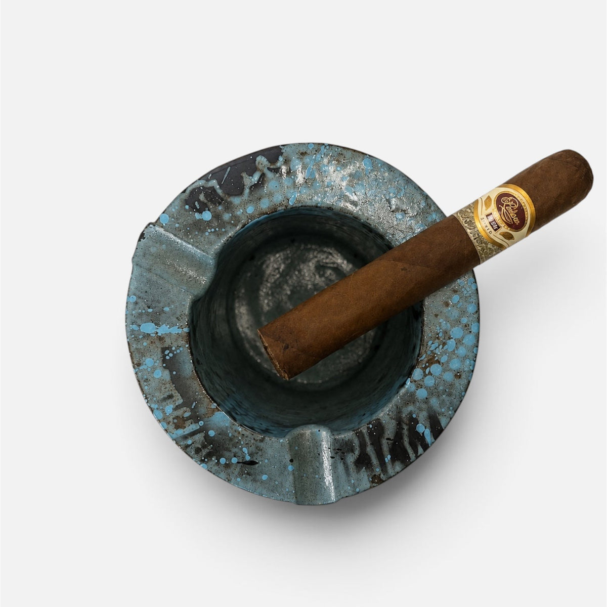 A brown cigar with a gold band resting diagonally inside a distressed blue and black ceramic ashtray, shot from a top-down perspective on a light gray background.