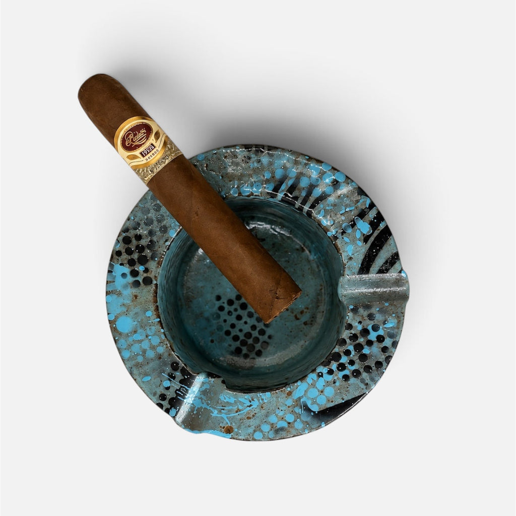 A single brown cigar with a gold and red band rests diagonally on a round, distressed blue and black patterned ceramic ashtray, photographed from a top-down perspective on a light gray background.