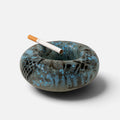A round ceramic ashtray with a textured, speckled glaze in shades of blue, gray, and black, shown on a light gray background with a single cigarette resting inside.