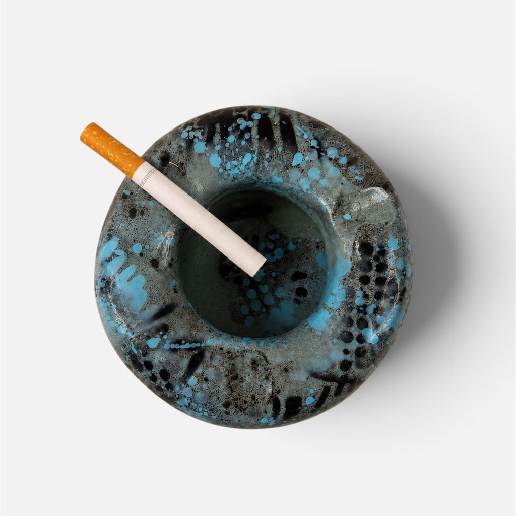 A round ceramic ashtray with a textured black and blue glaze, shown from a top-down view on a light gray background with a cigarette resting inside.