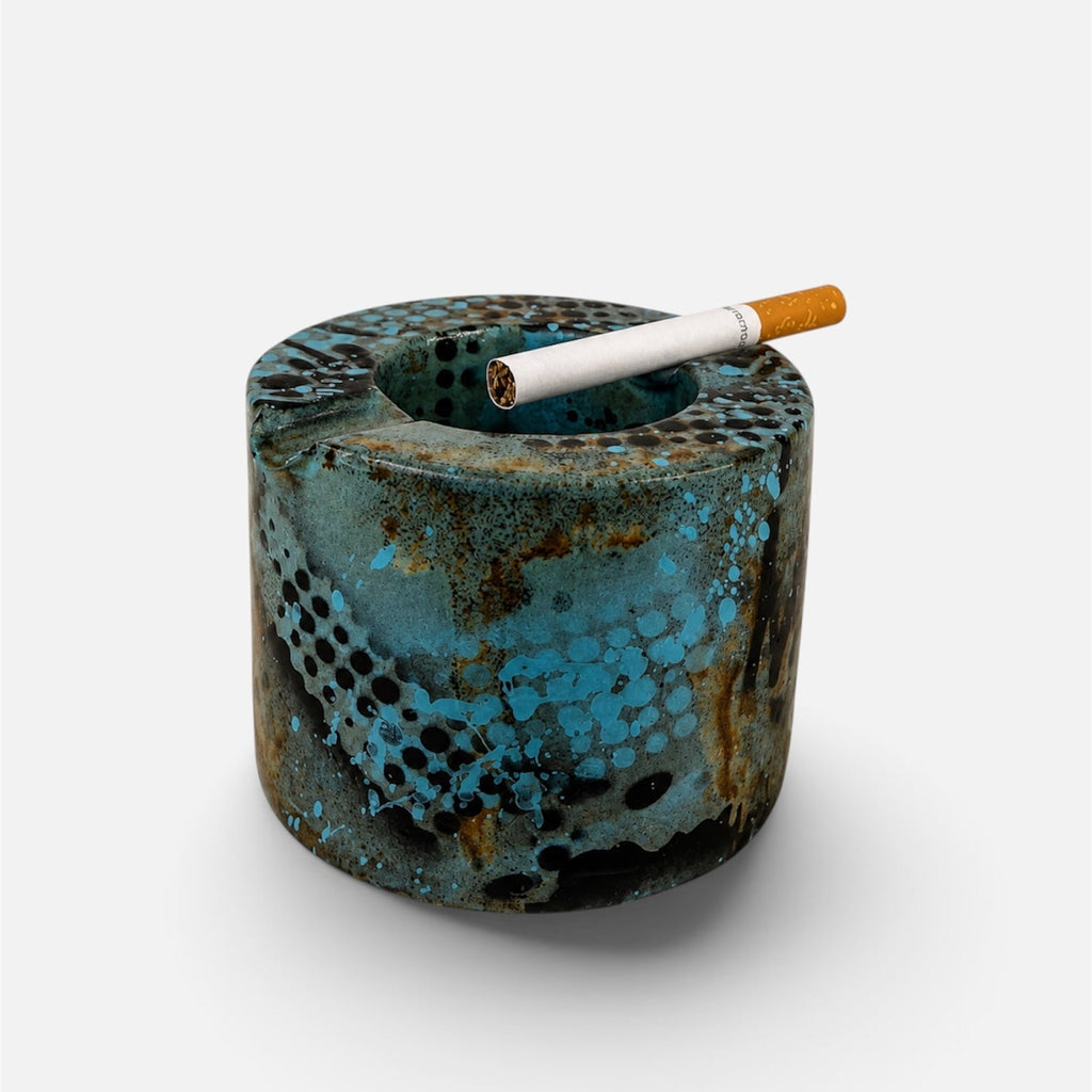 A cylindrical ceramic ashtray with a textured, distressed finish in turquoise blue, brown, and black, shown on a light gray background with a cigarette resting inside.