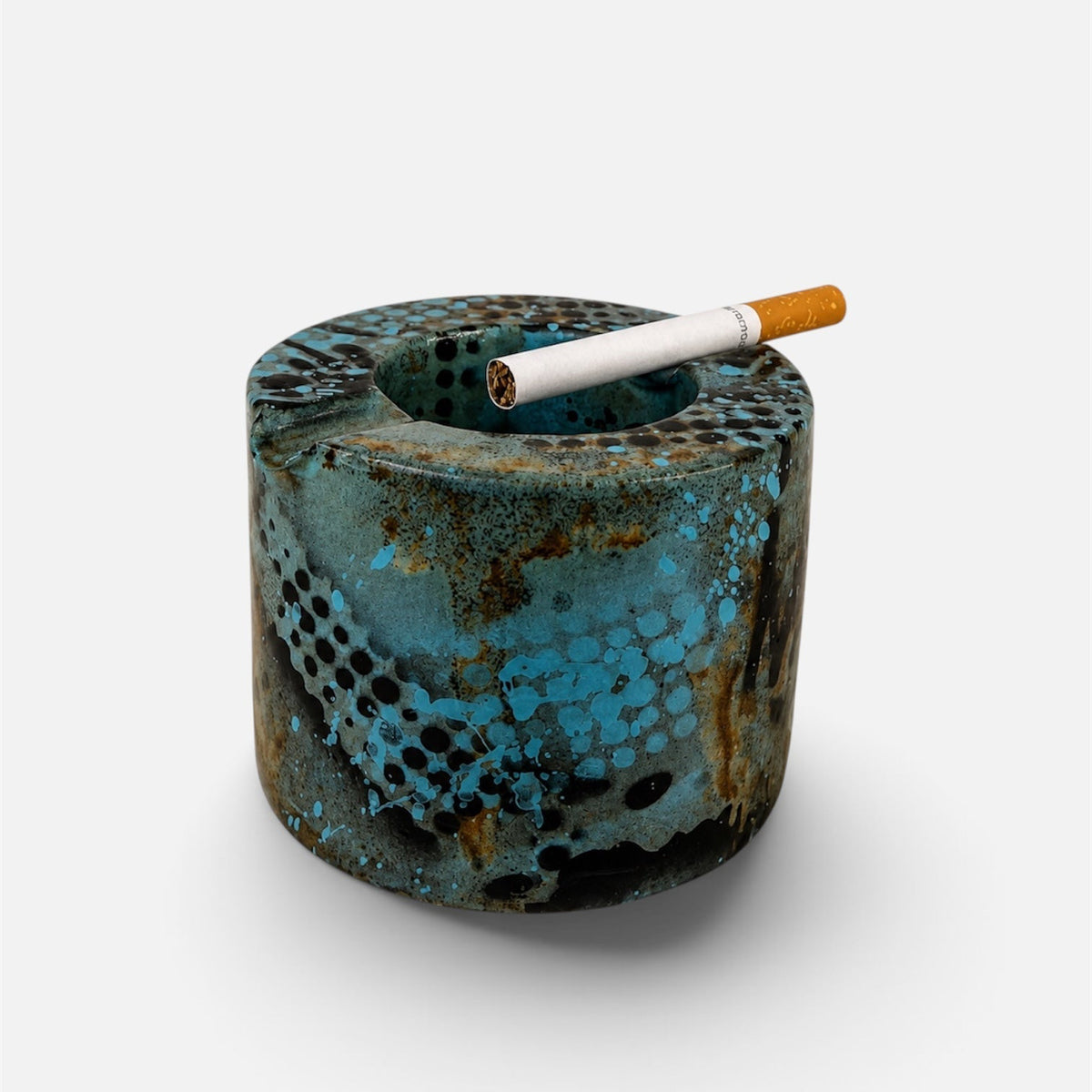 A cylindrical ceramic ashtray with a textured, distressed finish in turquoise blue, brown, and black, shown on a light gray background with a cigarette resting inside.