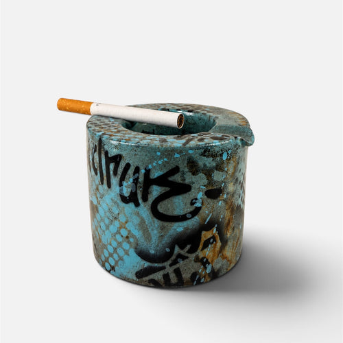 A cylindrical ceramic ashtray with a distressed turquoise blue and brown patina finish, featuring black graffiti-style lettering, with a cigarette resting across the top, set against a plain light gray background.