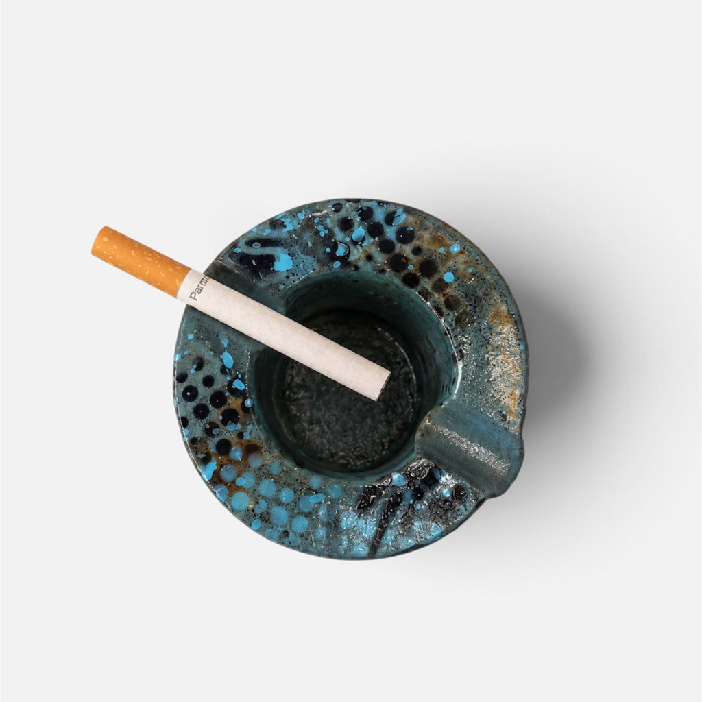 A top-down view of a round, speckled blue and black ceramic ashtray with a cigarette resting inside, set against a plain light gray background.