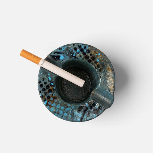 A top-down view of a round, speckled blue and black ceramic ashtray with a cigarette resting inside, set against a plain light gray background.