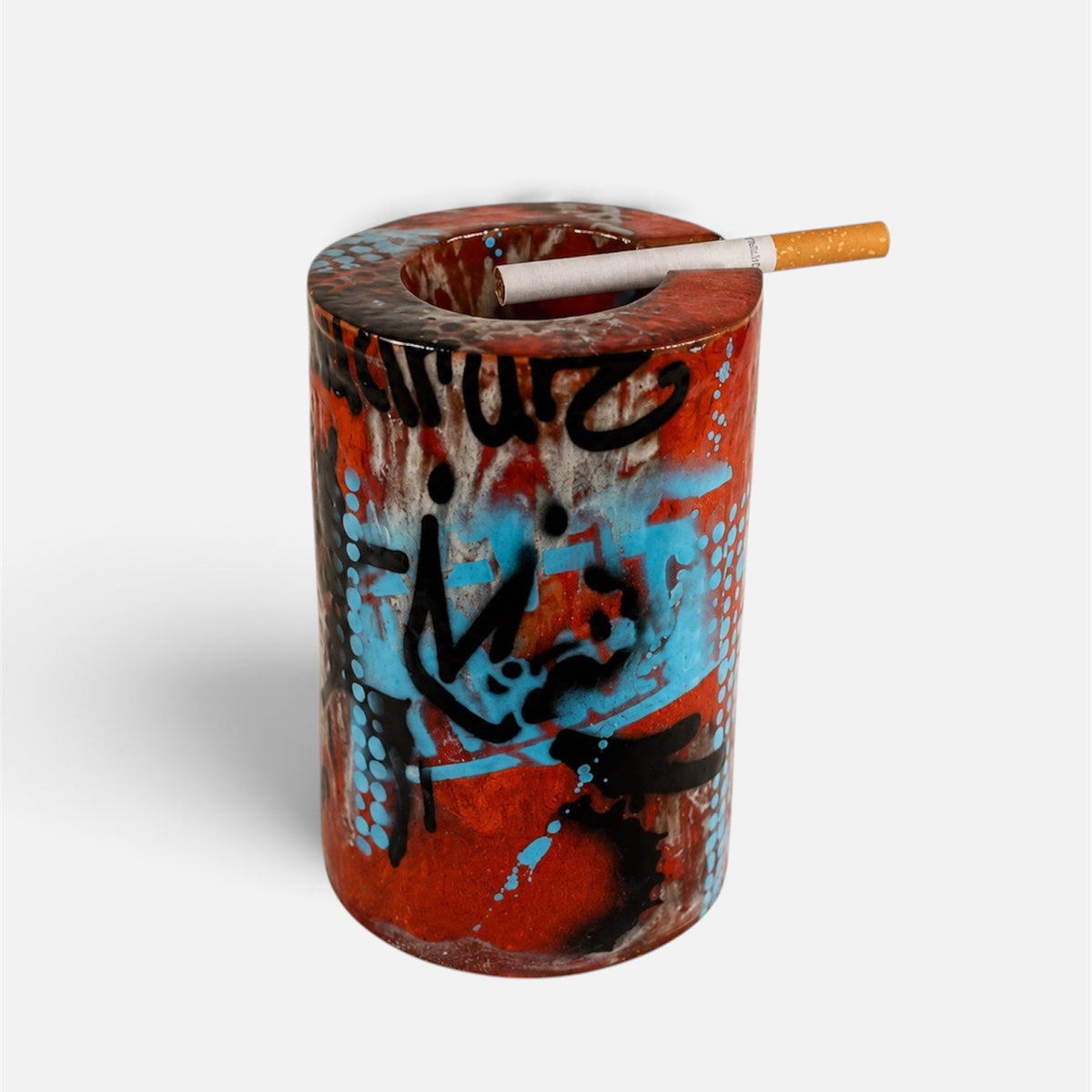 A cylindrical red and orange graffiti-style ashtray with blue and black spray-paint markings, shown on a light gray background with a cigarette resting across the top opening.