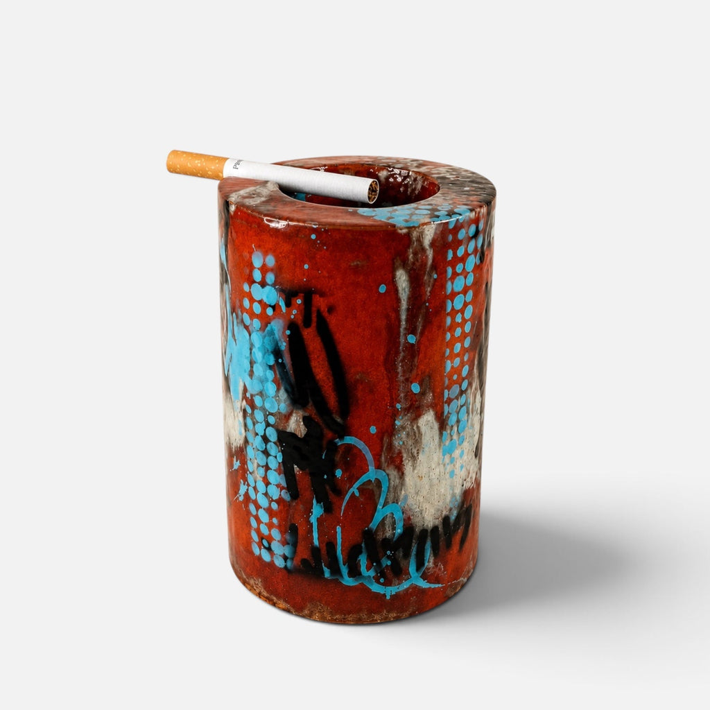 A cylindrical ceramic ashtray with a glossy red-brown glaze, decorated with blue, black, and white abstract patterns. A cigarette rests on top of the open container, which is photographed against a clean, light gray background.
