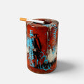 A cylindrical ceramic ashtray with a glossy red-brown glaze, decorated with blue, black, and white abstract patterns. A cigarette rests on top of the open container, which is photographed against a clean, light gray background.