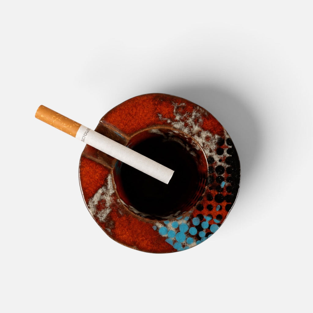 A top-down view of a round ceramic ashtray with a glossy red, orange, and blue pattern, featuring a lit cigarette resting inside the center on a clean white background.