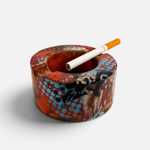 A round ceramic ashtray with a marbled pattern of orange, brown, black, and blue, featuring a white cigarette resting inside.