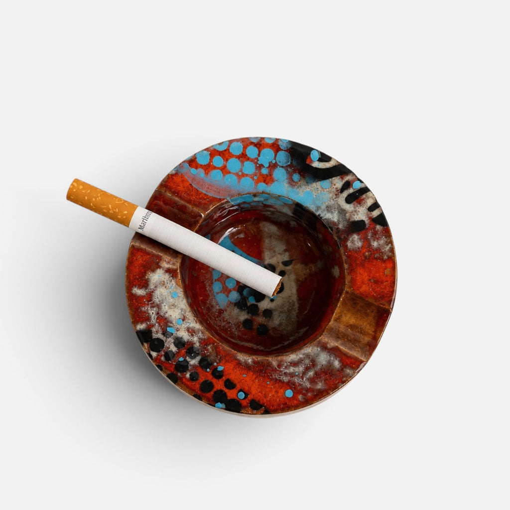 A top-down view of a round, hand-painted ceramic ashtray with a cigarette resting on it, set against a plain light gray background.