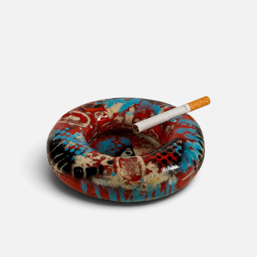 A circular ceramic ashtray with a colorful abstract pattern in red, blue, black, and beige, featuring a lit cigarette resting across the top, set against a plain light gray background.