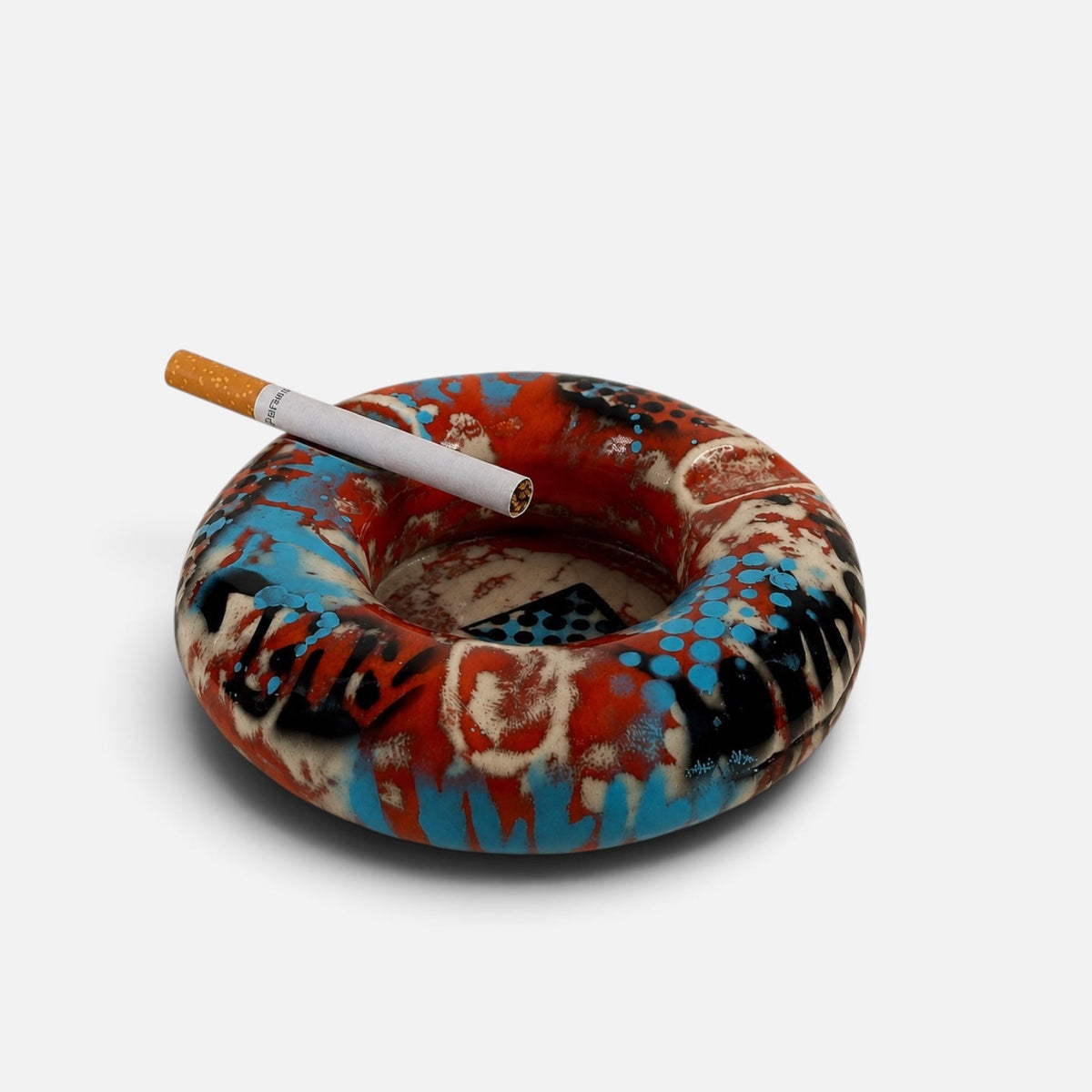 A circular glass ashtray with a colorful graffiti-style pattern in red, blue, black, and cream, shown on a light gray background with a cigarette resting on top.