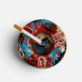 A round ceramic ashtray with an abstract graffiti-style pattern in red, blue, black, and beige, shown on a light gray background with a white cigarette resting inside.