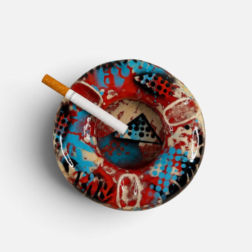 A round ceramic ashtray with an abstract graffiti-style pattern in red, blue, black, and beige, shown on a light gray background with a white cigarette resting inside.